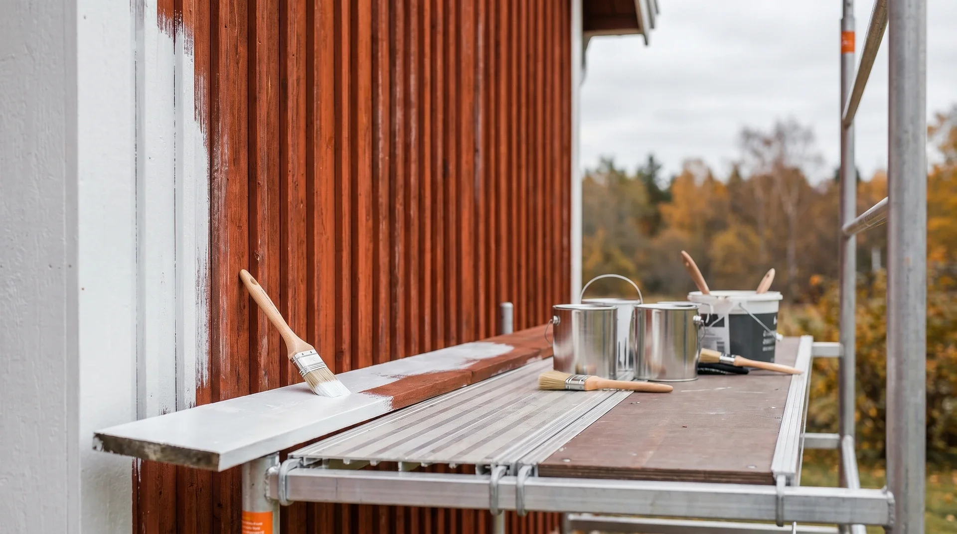 Målarfärg och penslar på ställning framför orangeröd trävägg med höstlandskap i bakgrunden.